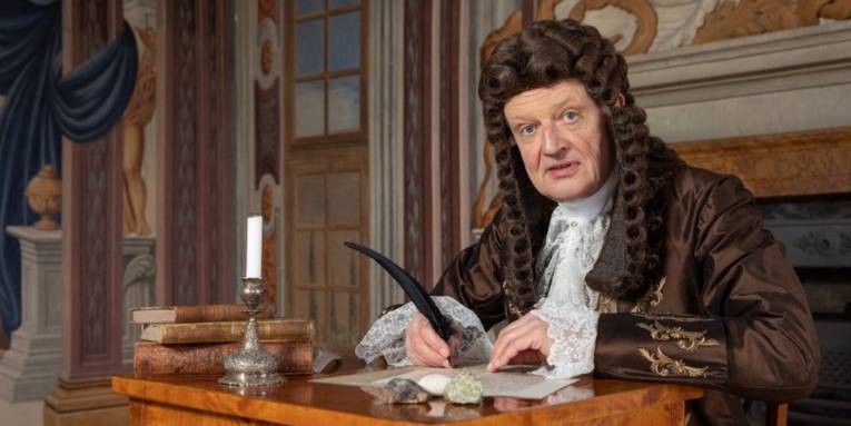 Mann mit Perücke in Kleidung aus der Barockzeit mit einer Schreibfeder in der Hand an einem Schreibtisch sitzend.